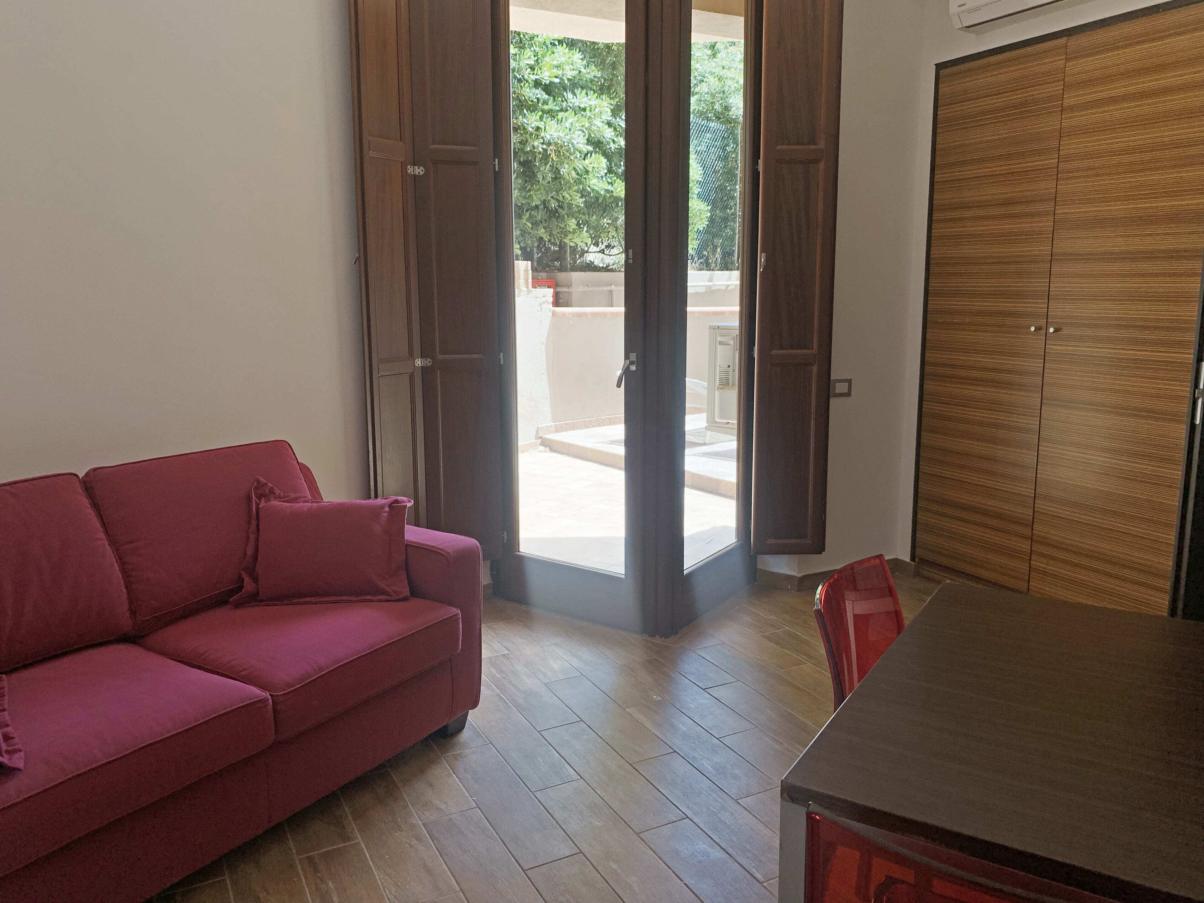 A room with a red sofa, a wooden wardrobe, and a sliding glass door leading to an outdoor area.