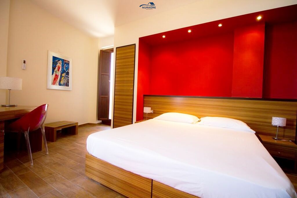 Quarto de hotel com uma cama grande, uma mesa com cadeira, uma parede de destaque vermelha e um quadro na parede.