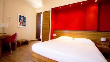 Quarto de hotel com uma cama grande, uma mesa com cadeira, uma parede de destaque vermelha e um quadro na parede.