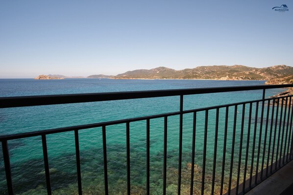 A coastal view from a balcony with clear turquoise water, a metal railing, and distant mountains.