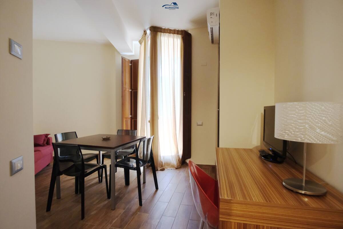 A dining area with a wooden table and chairs, a TV on a stand, and a window with curtains.
