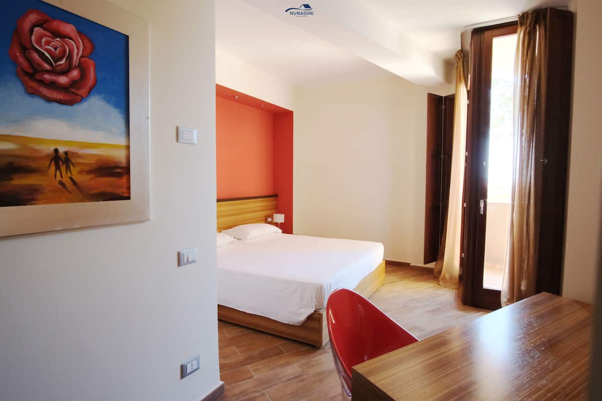 A hotel room with a bed, a desk, a chair, a painting of a rose, and a view of the outdoors.