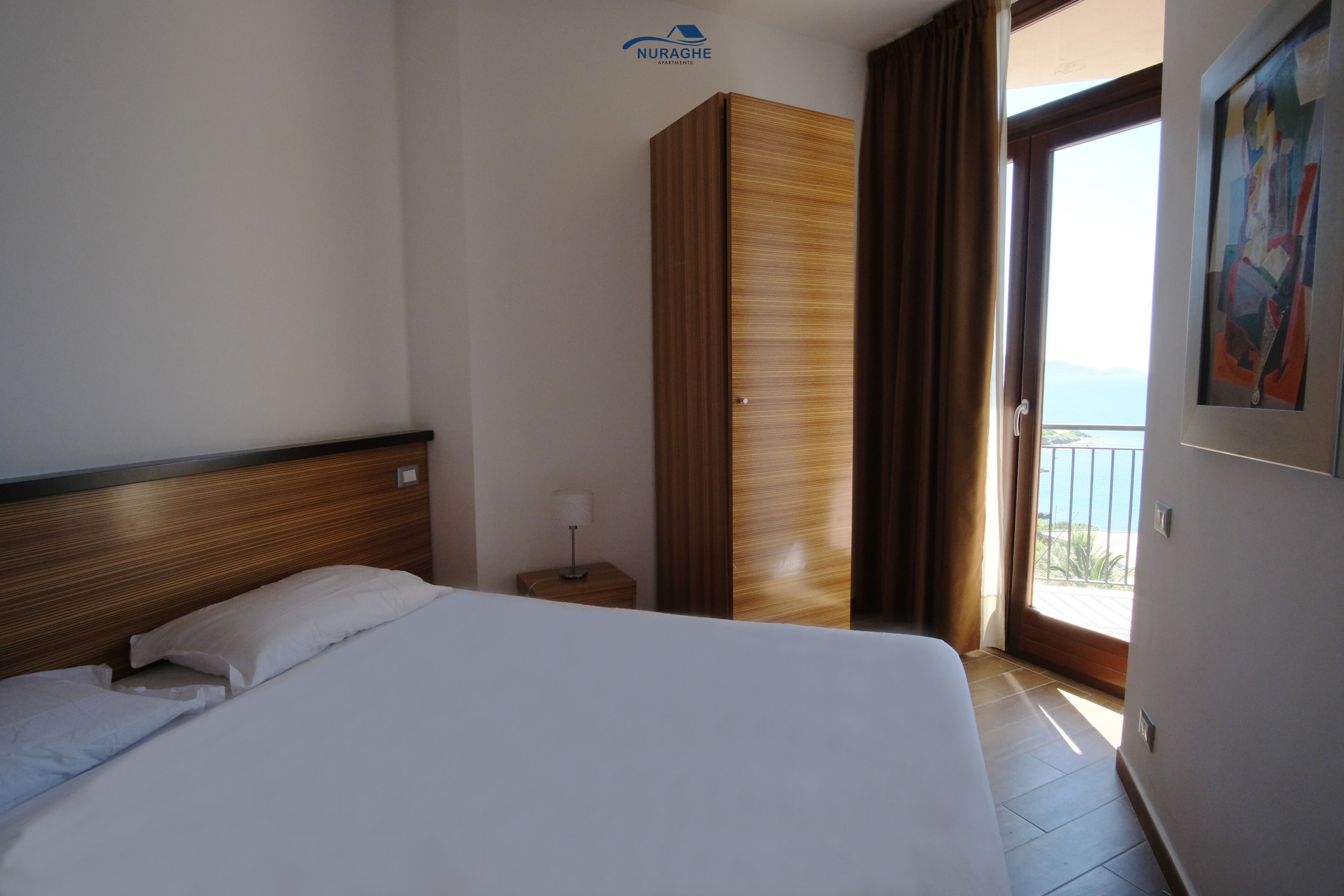 A hotel room with a bed, a wooden wardrobe, a nightstand with a lamp, and a balcony with a view.
