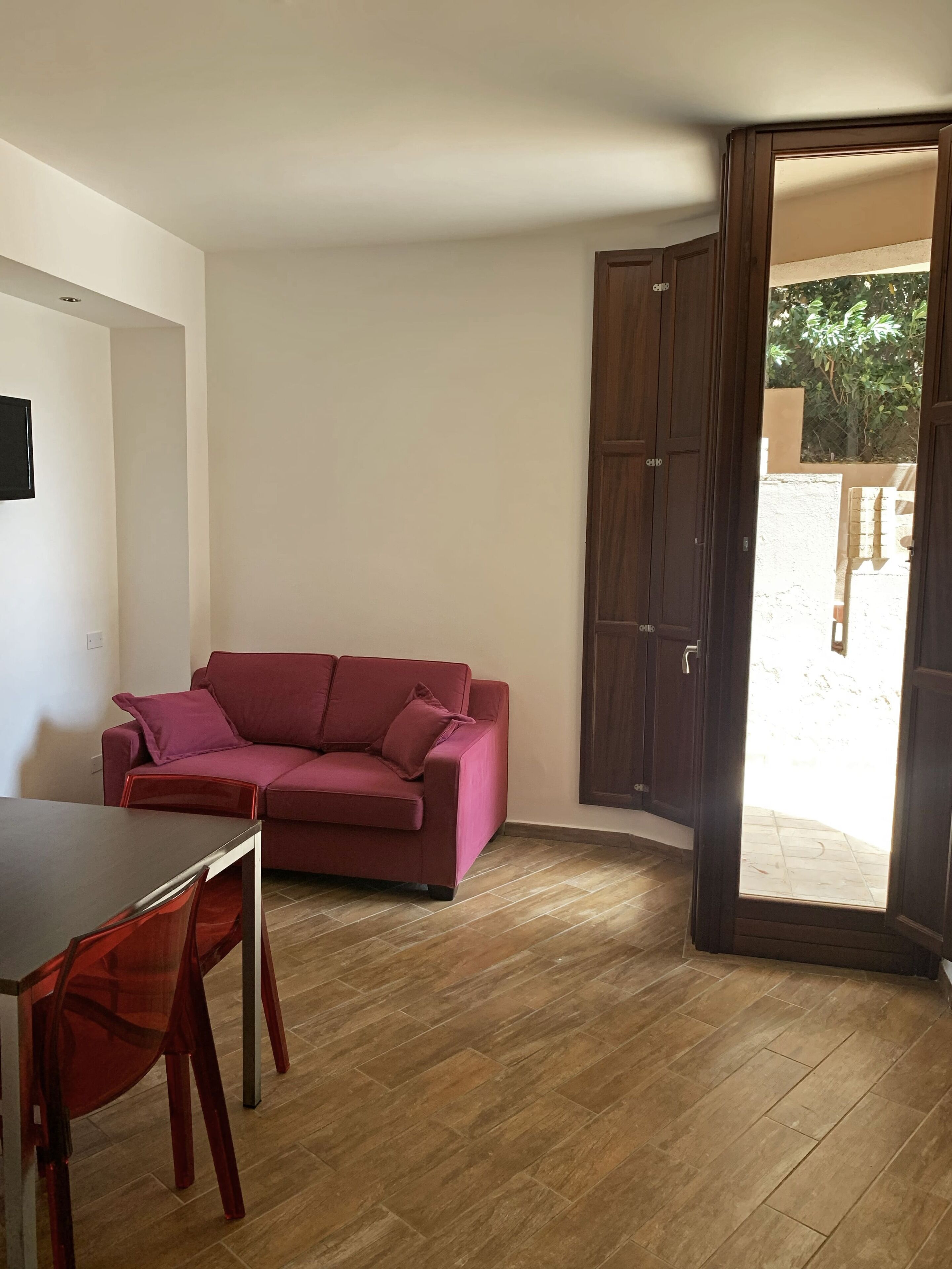 Ein Zimmer mit Holzboden, einem roten Sofa, einem kleinen Tisch und einer zur Außenwelt hinweisenden Tür.