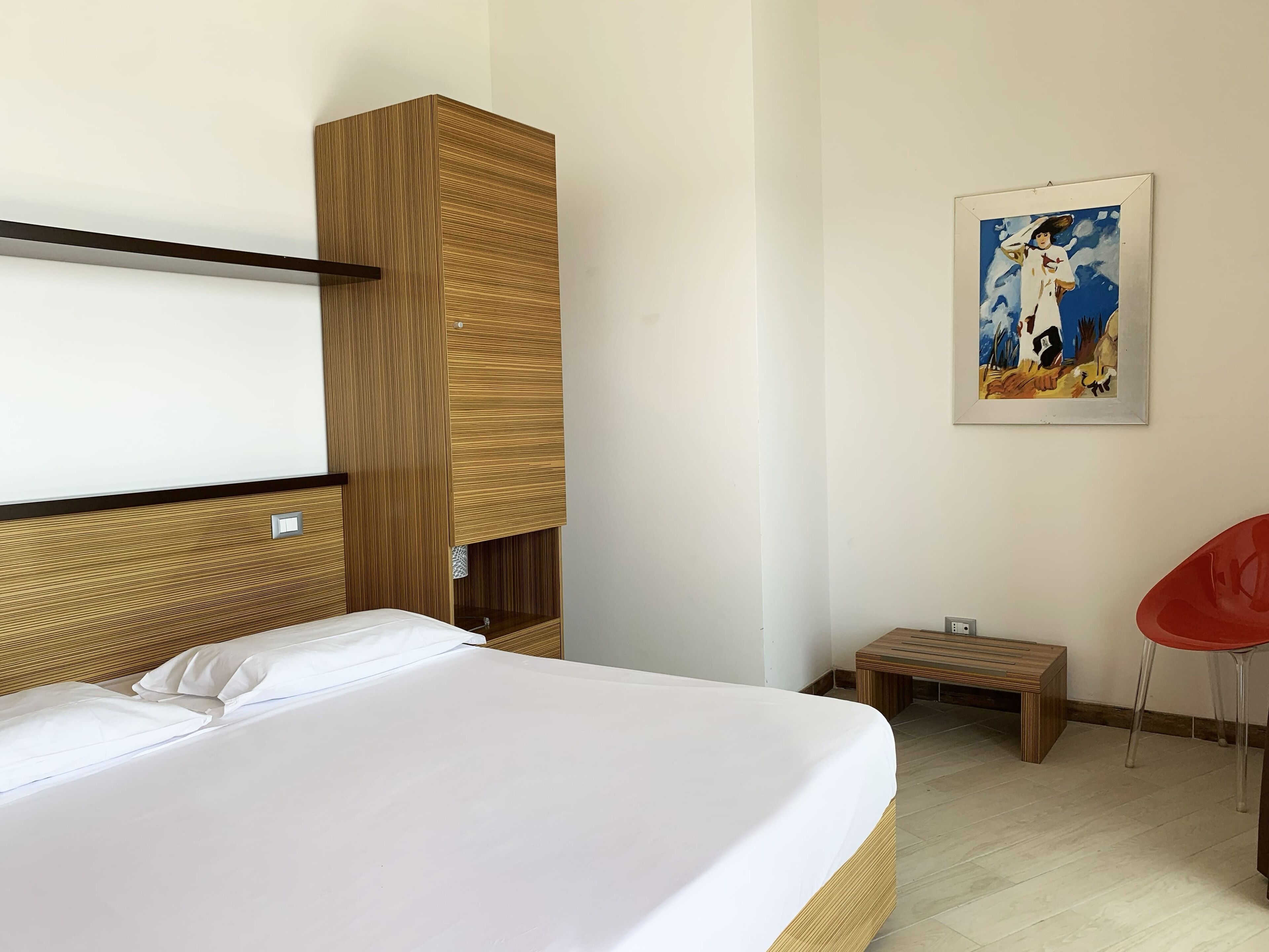 Ein Hotelzimmer mit Bett, einem Holzschrank, einem kleinen Tisch, einem roten Stuhl und einem Bild an der Wand.