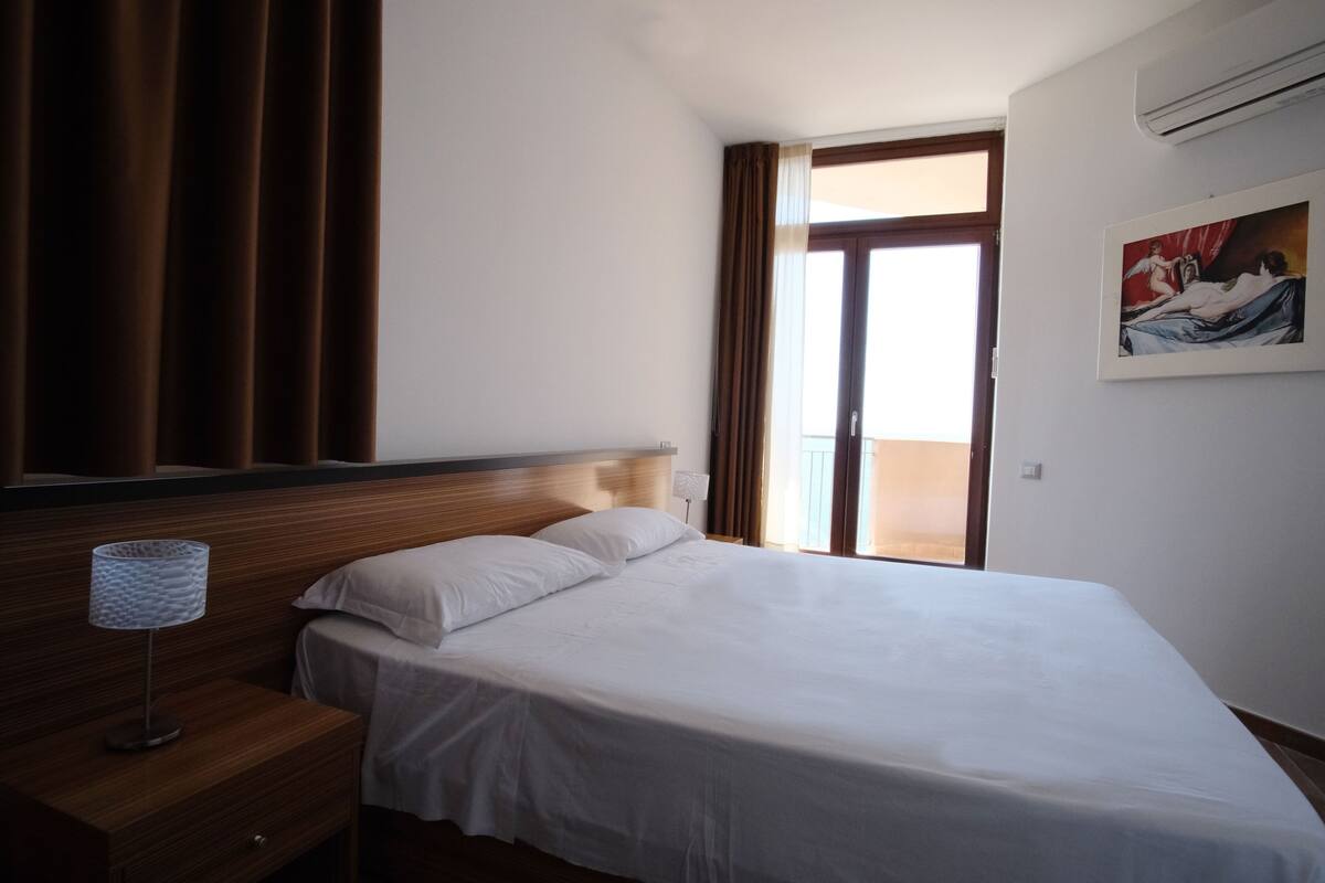 A hotel room with a bed, a nightstand with a lamp, a window with curtains, and a painting on the wall.