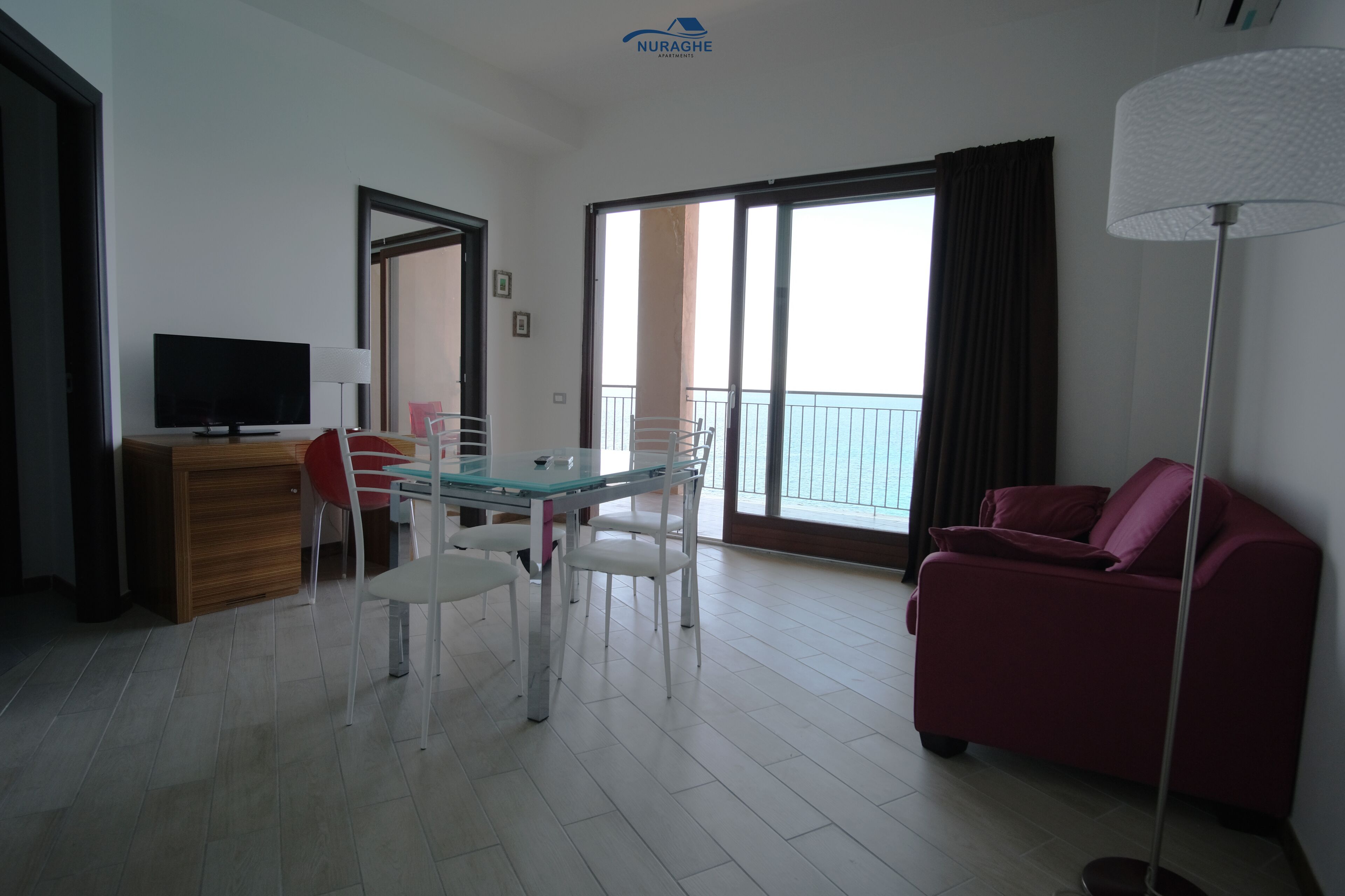 A modern living room with a glass dining table, white chairs, a red sofa, a flat-screen TV, and a balcony with a view.