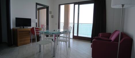 A modern living room with a glass dining table, white chairs, a red sofa, a flat-screen TV, and a balcony with a view.