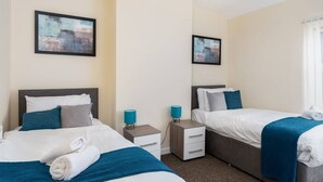 A hotel room with two beds, each with a blue blanket and pillows, a nightstand with a lamp, and a framed picture on the wall. - Cecil Lodge - 2 Bed Apartment (Swansea)