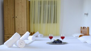 Een hotelkamer met een netjes opgemaakt bed, twee opgerolde handdoeken, een dienblad met druiven en twee glazen rode wijn, en een houten garderobe.