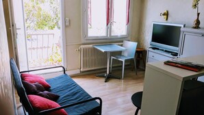 A small, well-lit room with a bed, a desk with a computer, a chair, and a TV.