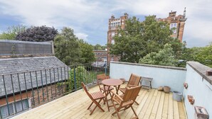 A wooden deck with a table and chairs, a potted plant, and a brick wall. - JOIVY Charming 3 bed house with a rooftop terrace (London)
