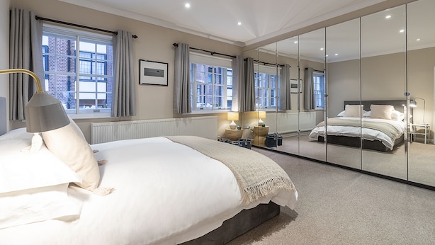 A hotel bedroom with a large bed, a bedside table, a lamp, a mirror, and windows with curtains. - JOIVY Stunning 3BR house near Regents Park& Baker Street (London)