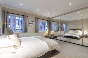 A modern bedroom with a large bed, a bedside table with lamps, a mirror, and a view of the outside through large windows.