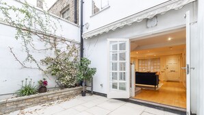 A residential property with a white exterior, a garden with plants and a tree, and an open door leading to a room with a black sofa and wooden flooring. - JOIVY Stunning 3BR house near Regents Park& Baker Street (London)