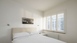 A bedroom with a bed, a nightstand, a wall-mounted picture, and a window with blinds. - JOIVY Sublime 1 bed flat with Thames view (London)