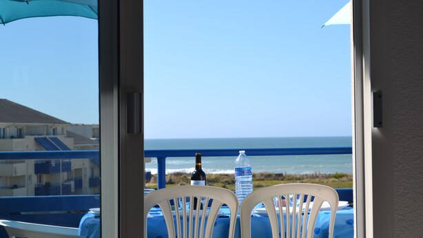 A balcony with a table and chairs, a bottle of water, and a bottle of wine, overlooking the ocean and buildings. - Waterfront apartment (Lacanau Océan)