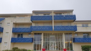 A multi-story building with blue balconies, a central entrance with glass doors, and a small garden area with potted plants. - Waterfront apartment (Lacanau Océan)