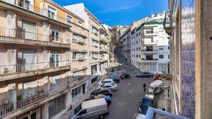 Una estrecha calle urbana con edificios de varios pisos, coches estacionados y un cielo azul despejado.