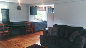 Sala de estar com sofá escuro, piso de madeira, área de cozinha com janela e um relógio na parede.