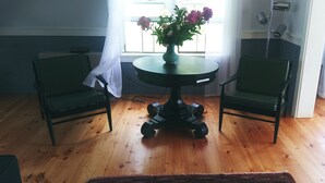Quarto com piso de madeira, uma mesa redonda com um vaso de flores, duas cadeiras verdes e uma janela com cortinas.