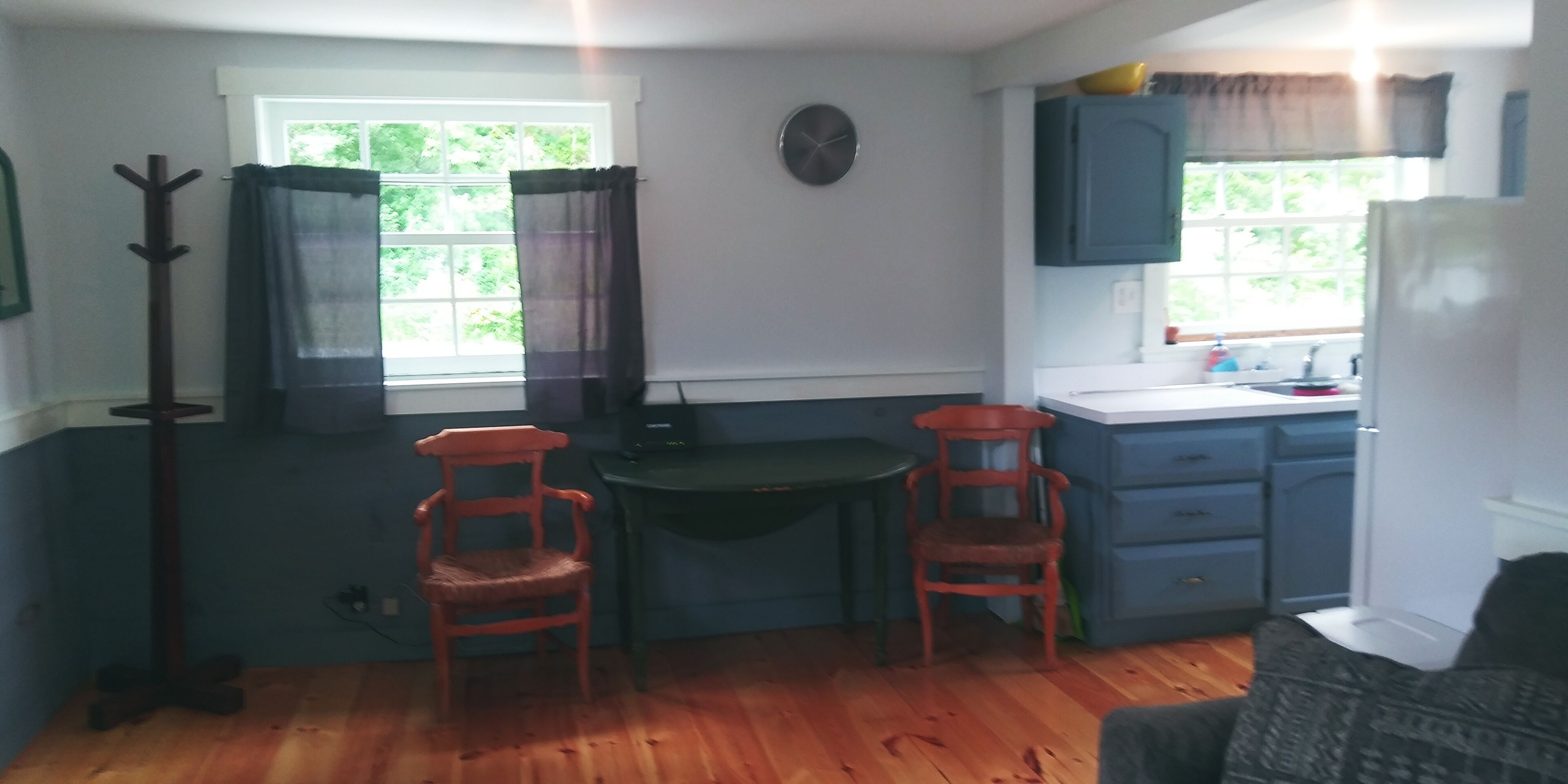 A kitchen with wooden flooring, a window with curtains, a clock on the wall, and a sink area.