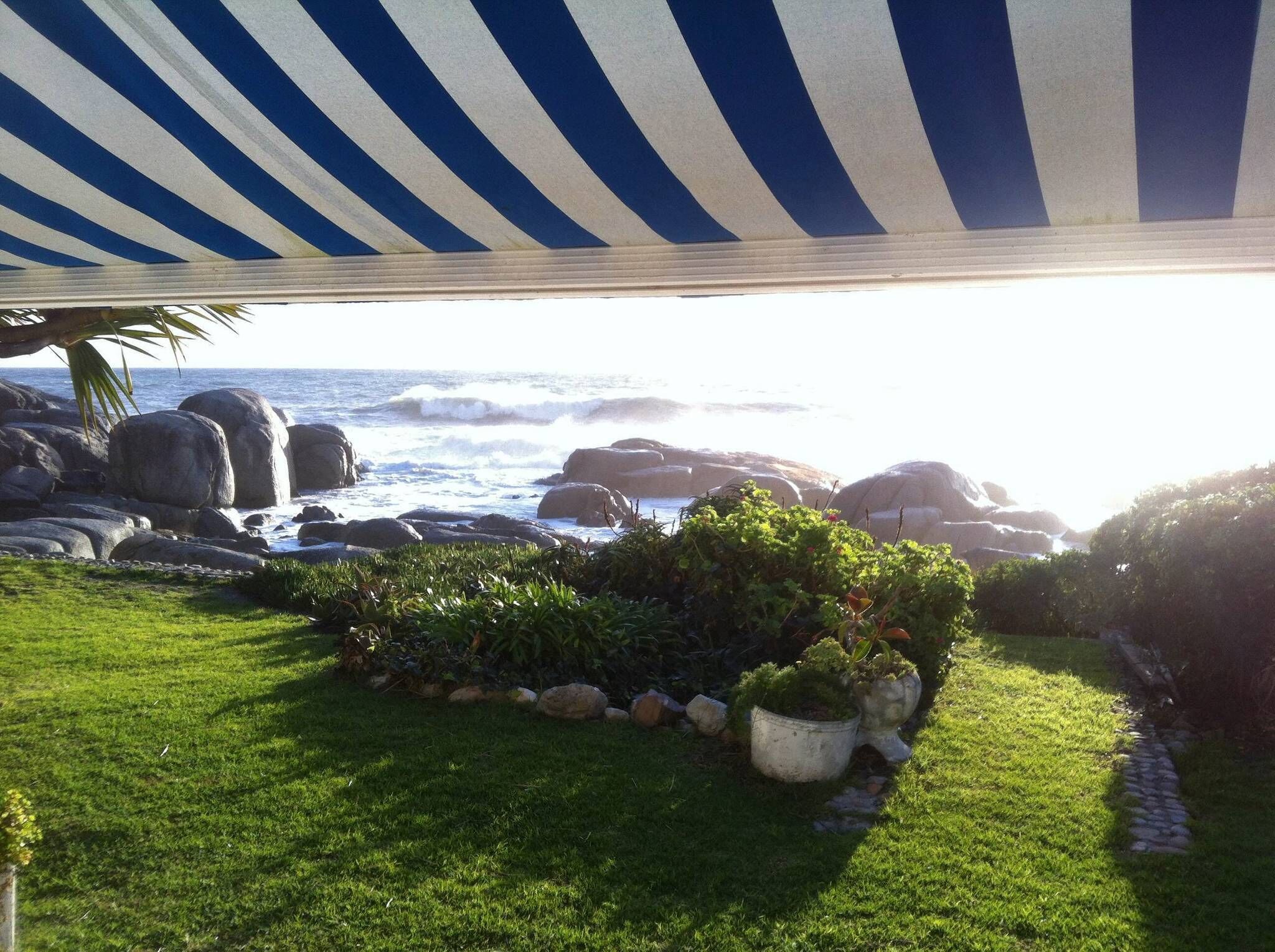 A coastal garden with a view of the ocean, featuring a striped awning, lush greenery, and large rocks.