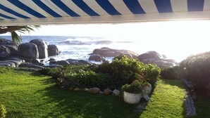 Un jardín costero con vista al océano, que cuenta con un toldo a rayas, abundante vegetación y grandes rocas.