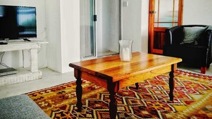 Una sala de estar con un televisor sobre un soporte blanco, una mesa de madera con un florero, un tapiz estampado y una silla negra.