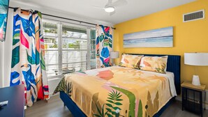 A bedroom with a bed, a ceiling fan, a window with blinds, a painting on the wall, and a nightstand with a lamp. - Walk to the Beach | Osprey Villa | Siesta Key (Sarasota)
