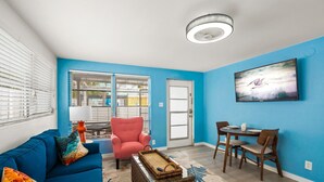 A living room with a blue sofa, a pink armchair, a dining table with chairs, and a television on the wall. - Walk to the Beach | Osprey Villa | Siesta Key (Sarasota)