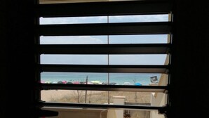 A view through window blinds to a beach with umbrellas and a clear sky.