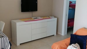 A bedroom with a bed, a TV mounted on the wall, a bedside table, and a bunk bed in the background.