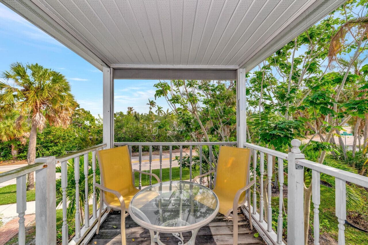 A covered outdoor area with a glass table and two yellow chairs, surrounded by white railings and lush greenery.