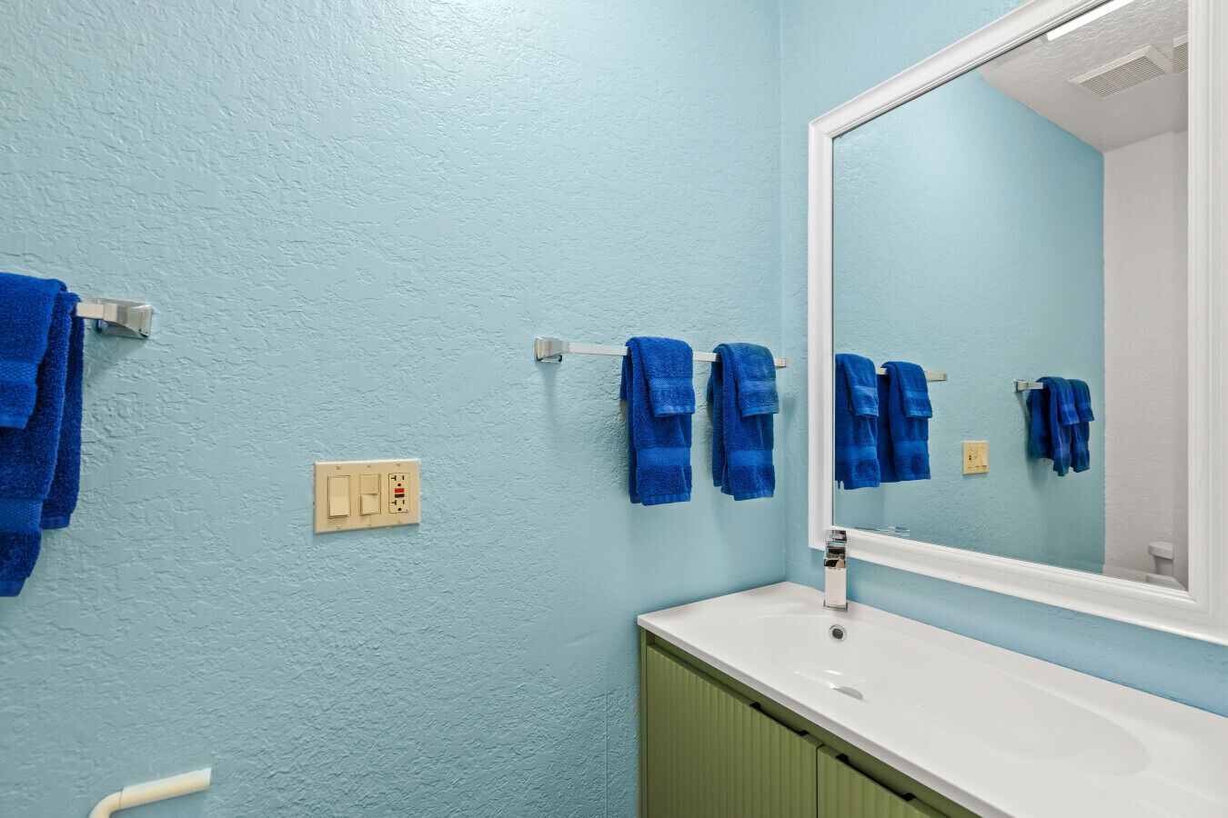 A bathroom with a light blue wall, a white sink, and a mirror. There are multiple blue towels hanging on a rack.