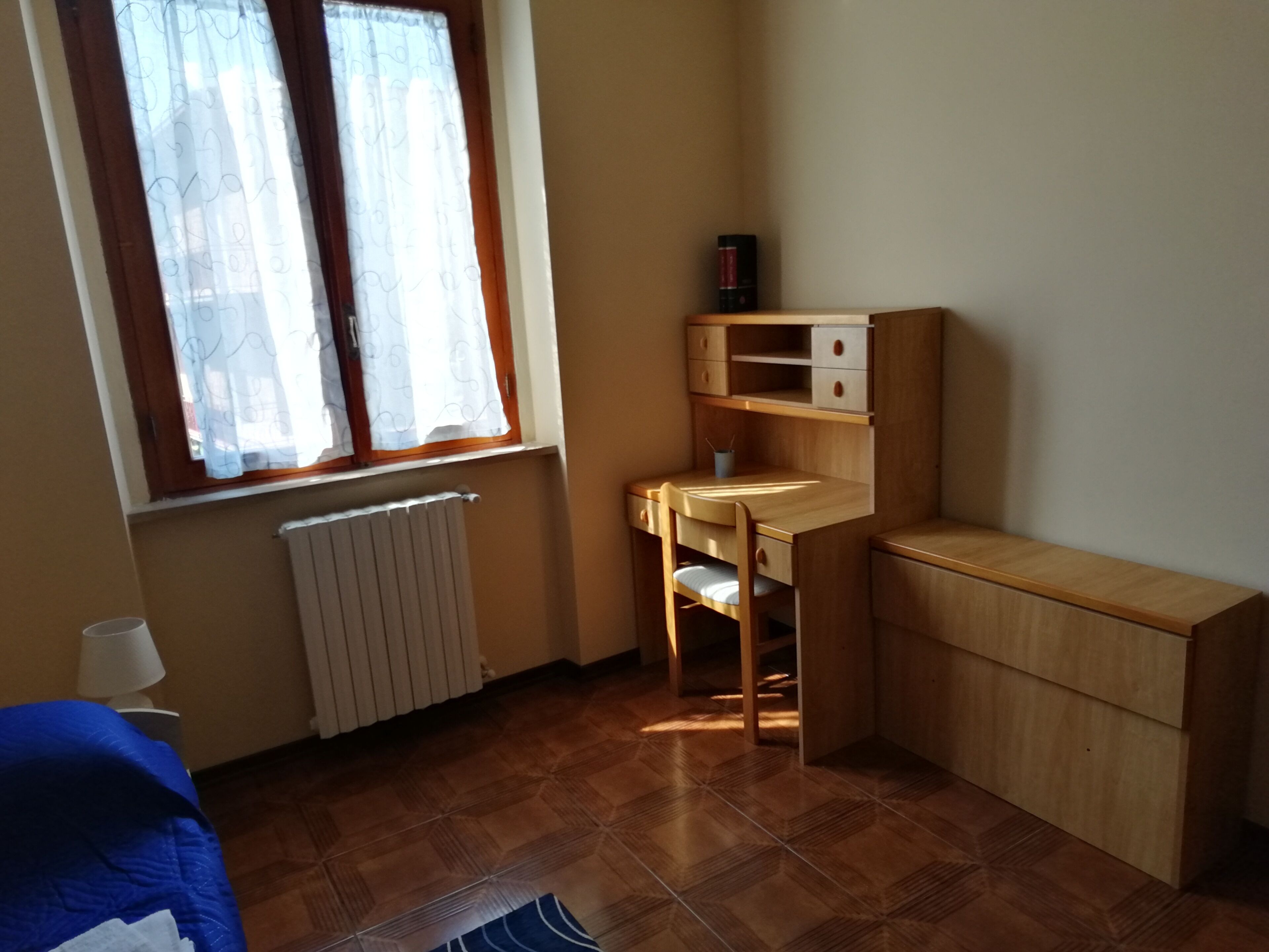 A small, well-lit room with a wooden desk, a chair, a radiator, and a window with curtains.