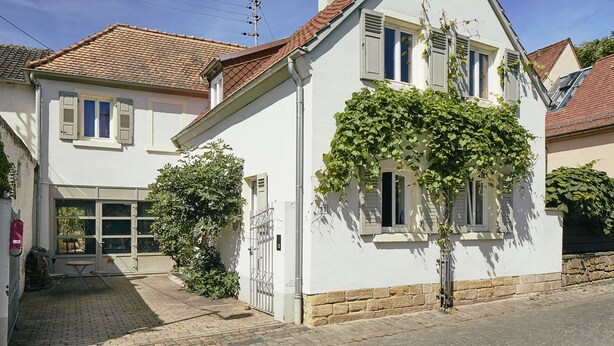 A two-story house with a tiled roof, white walls, and shutters. There is a small garden area with a gate and a tree.