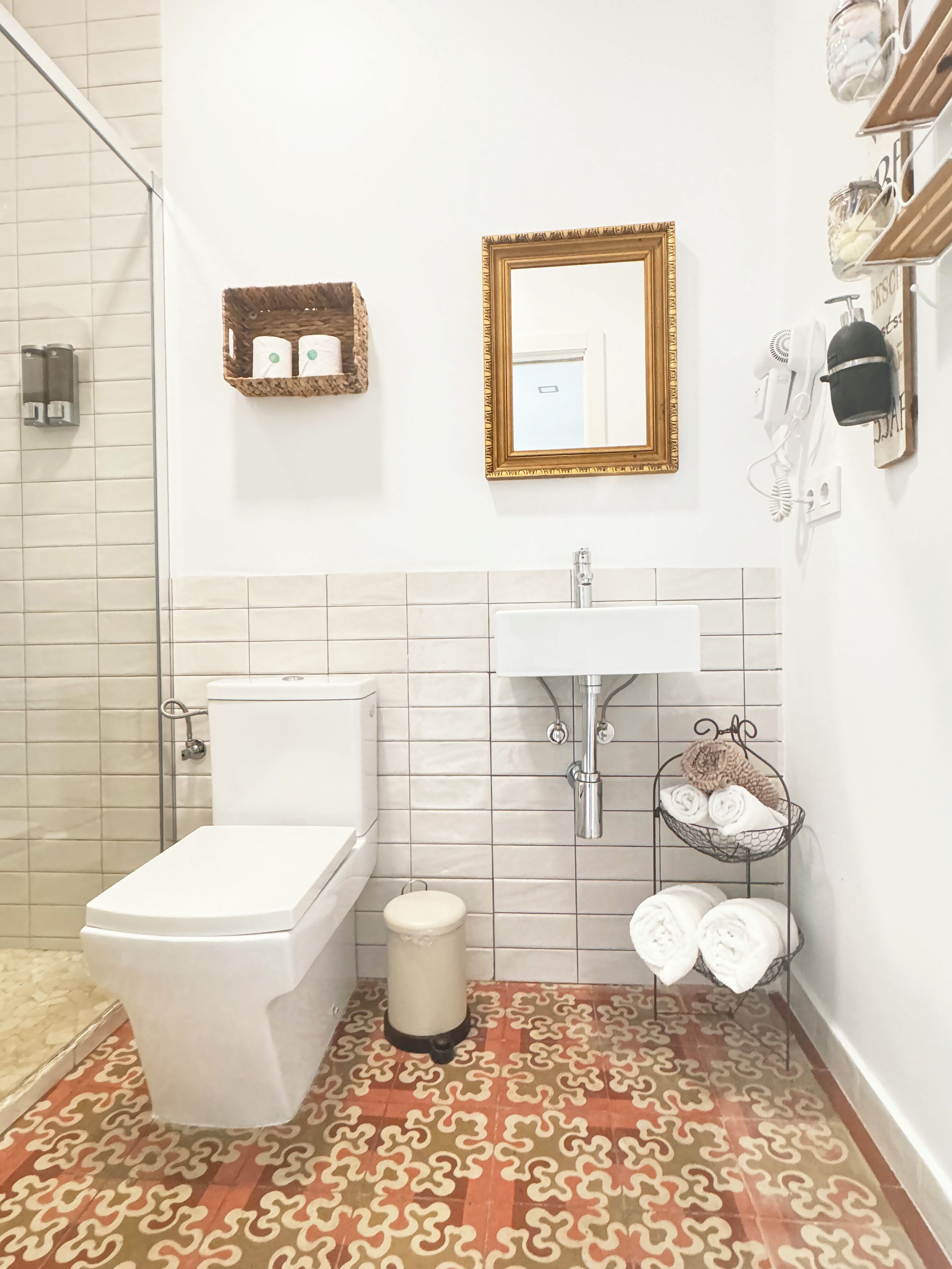 A bathroom with a toilet, sink, and shower. There is a patterned rug on the floor and a small trash bin.