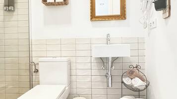 A bathroom with a toilet, sink, and shower. There is a patterned rug on the floor and a small trash bin.