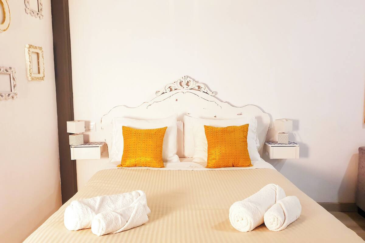 A neatly made bed with white and yellow pillows, a striped bedspread, and two rolled towels on the bed.