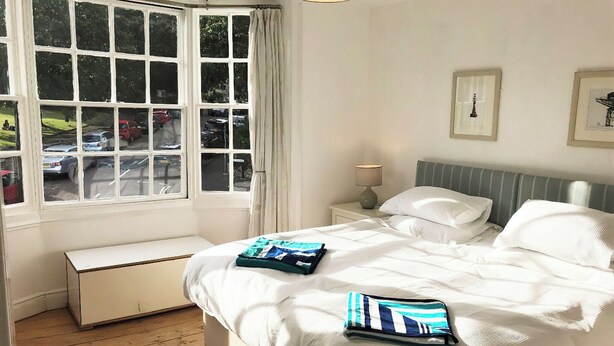 Ein Schlafzimmer mit großem Fenster, einem Nachttisch mit Lampe, einem Bett mit Kissen und einer Sitzbank am Fußende.
