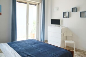 Une chambre à coucher avec un lit, un téléviseur posé sur une commode, une chaise et un balcon avec des rideaux.