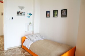 A single bed with an orange frame and a beige bedspread, a white wall with three framed pictures, a small white shelf with a plant, and a stone-tiled floor.
