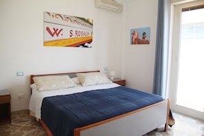 A bedroom with a bed, a nightstand, a wall-mounted air conditioner, a window with curtains, and a painting.