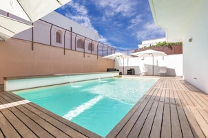 Una piscina con cubierta de madera, sombrillas blancas y un edificio blanco con ventanas arqueadas.