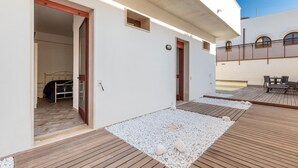 Un espace extérieur moderne doté d’une terrasse en bois, d’un petit coin salon et d’une chambre visible à travers une porte ouverte.