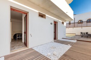 Un espacio exterior moderno con una terraza de madera, una pequeña zona de estar, y un dormitorio visible a través de una puerta abierta.