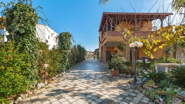 A paved pathway leading to a building with a balcony, surrounded by lush greenery and potted plants. - Villa Rosy (Maruggio)