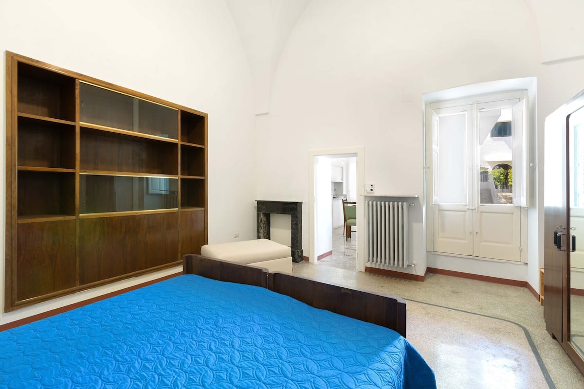 A bedroom with a large wooden wardrobe, a bed with a blue quilt, a small bench, and a radiator.