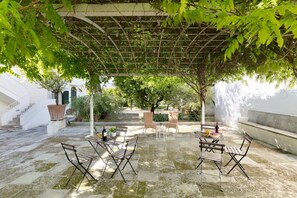 Un patio exterior con suelo de piedra, sillas de metal y una pérgola cubierta de plantas trepadoras.